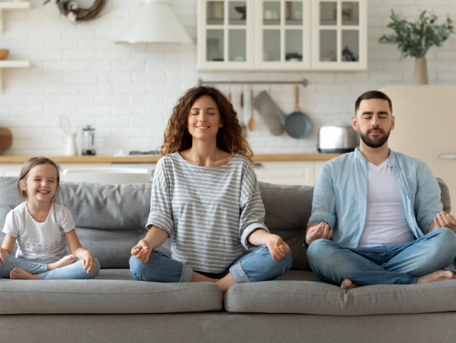 Calm young family with little daughter sit on couch practice yoga together, happy parents with small preschooler girl child rest on sofa meditate relieve negative emotions on weekend at home