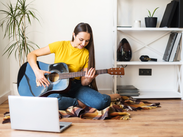 Hobbies and leisure activities during quarantine. Online training, online classes. A young woman watches a video lesson on playing the guitar, she sits on a cozy plaid with a guitar
