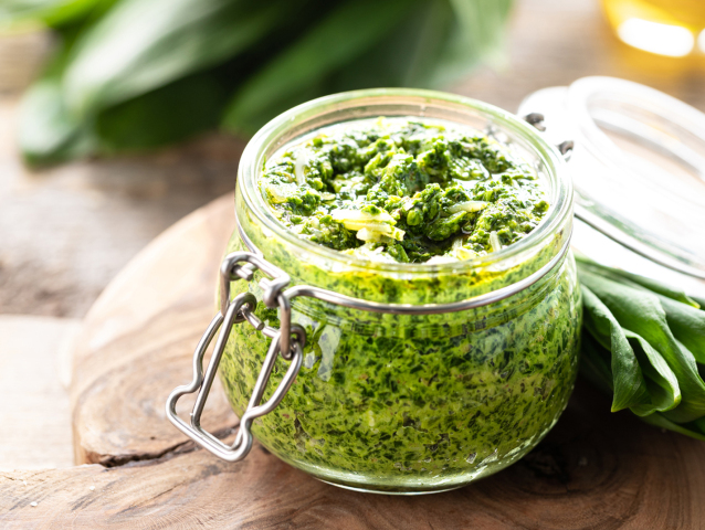 Wild leek pesto with olive oil and parmesan cheese in a glass jar on a wooden table. Useful properties of ramson. Leaves of fresh ramson. Horizontal