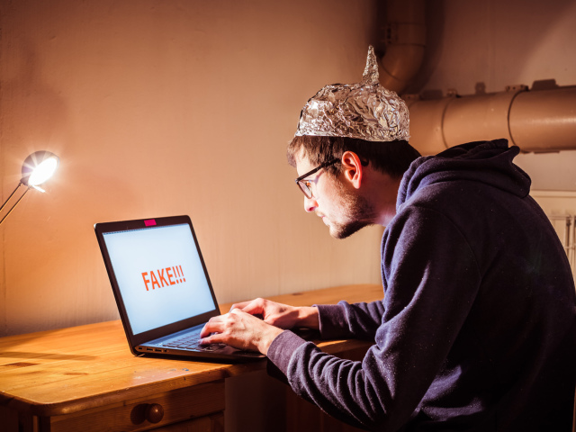 Young man with aluminum cap is sitting in the dark basement in front of a laptop. Conspiracy theory concept